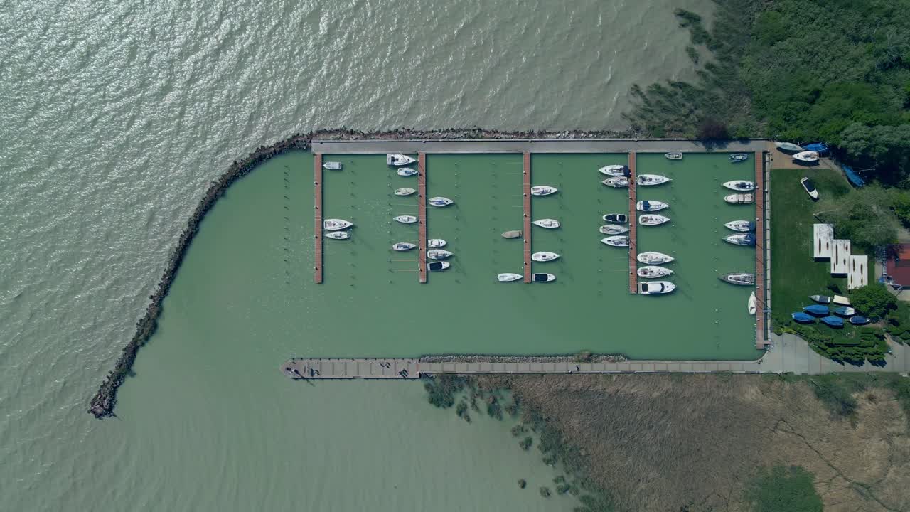 A top-down aerial view of a marina with multiple docks and moored boats, set in striking turquoise water. The harbor is bordered by a breakwater and natural shoreline, with green vegetation.