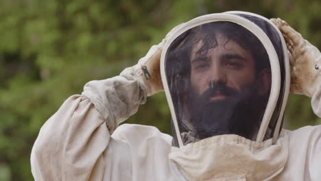 bearded asian beekeeper puts on hood of white protective bee suit, frontal slomo