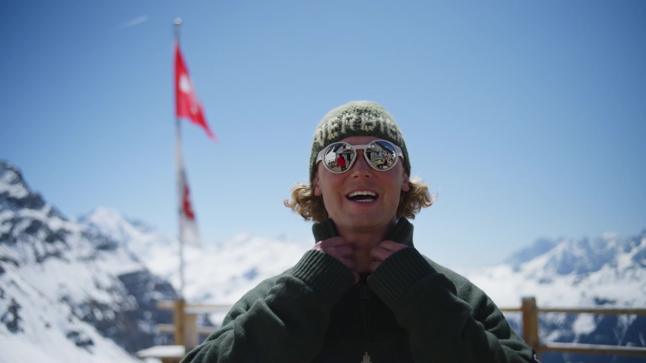 Giovane uomo con occhiali da sole e abbigliamento invernale che si gode viste panoramiche sulle Alpi svizzere a Verbier, Svizzera. Stile di vita alpino rilassato in una destinazione montana di lusso in una soleggiata giornata di primavera.