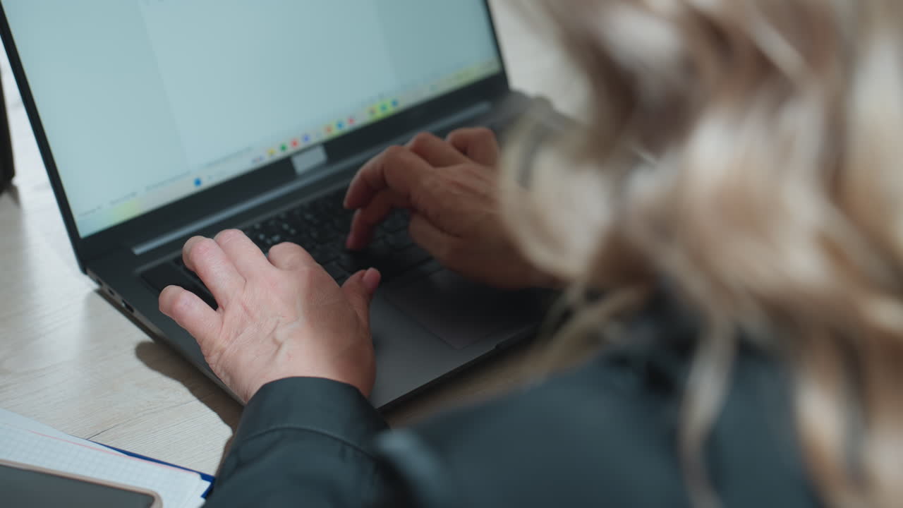 Close up rear view of woman typing on laptop at wooden desk in modern office, focused hands on keyboard and soft waves of blonde hair in foreground