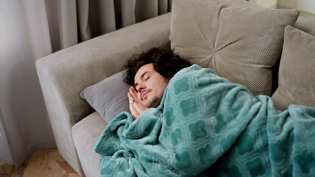 tranquilo dormido chico moreno con cabello rizado se cubrió con una manta azul y duerme en el sofá en un apartamento moderno