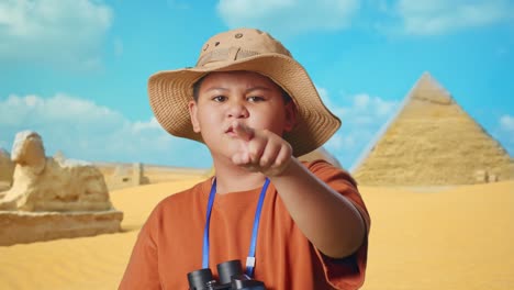 asian boy with a hat and binoculars pointing his finger towards you and furiously speaking against blaming you while traveling in giza pyramid. boy researcher, travel adventure, close up