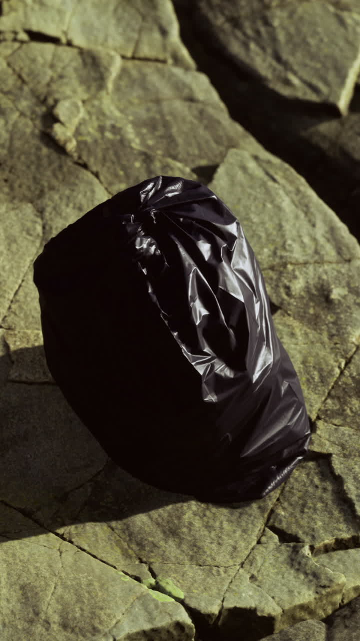 Black trash bag rests on rugged stones under the afternoon sun by the riverbank