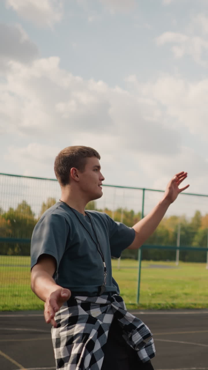entrenador con silbato alrededor del cuello golpea el voleibol con camisa a cuadros atada alrededor de la cintura, en una arena deportiva al aire libre con equipos deportivos en el fondo