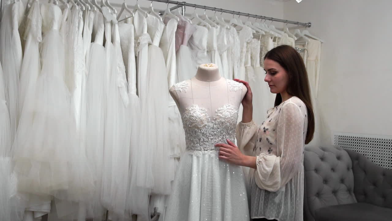 다른 드레스와 비교하여 객실에서 아름다운 미소의 웨딩 드레스를 찾고 있습니다. 결혼식을 준비하고 있습니다. 웨딩 드레스를 구입하고 있습니다. 바느질은 완성된 맞춤형 웨딩 드레스를 검사합니다.