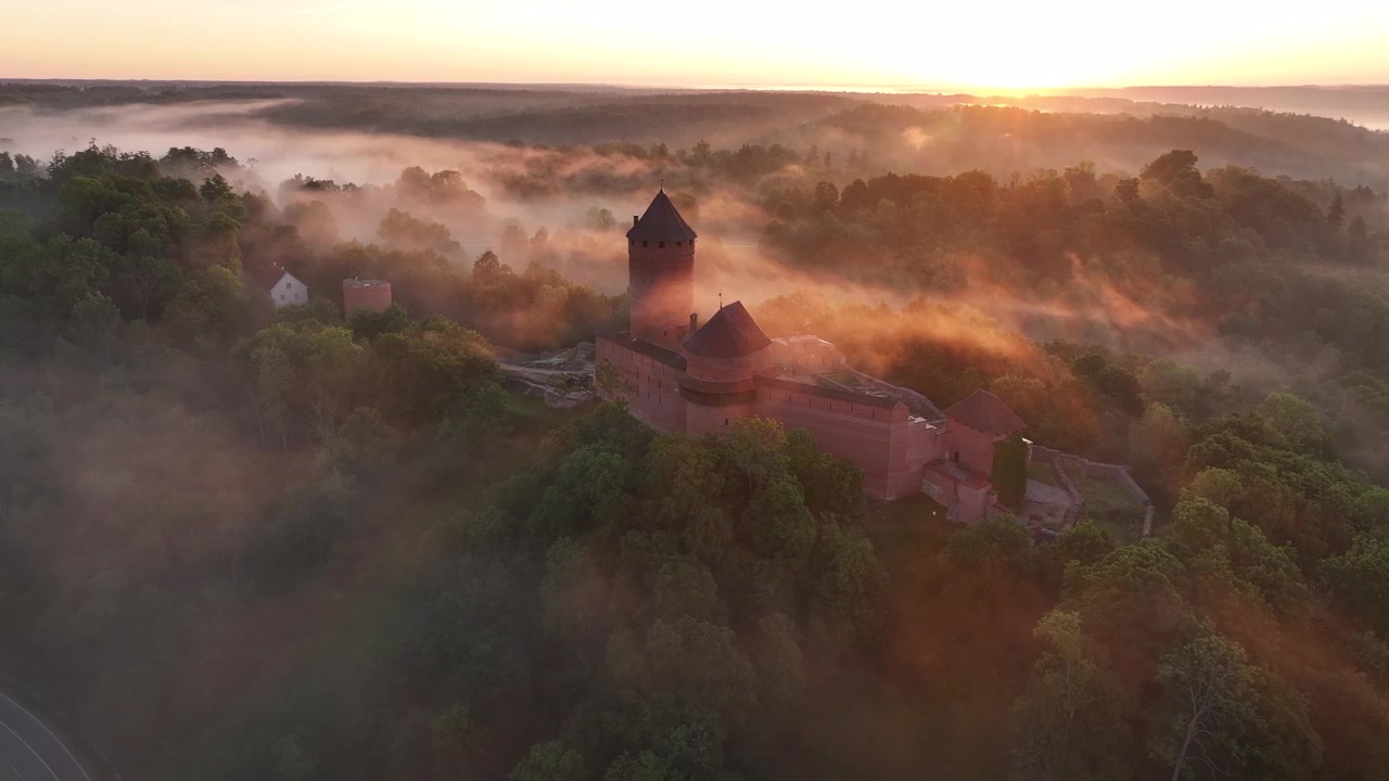 Cinematic drone footage of Turaida Castle at sunrise in Sigulda, Latvia. Golden sunlight illuminates the medieval red brick fortress while morning mist drifts through the Gauja River valley.