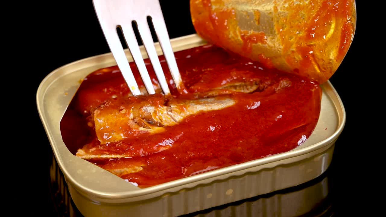 A metal fork lifts a sardine from an open tin can filled with tomato sauce, under bright studio lighting and against a black background
