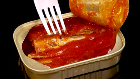 a metal fork lifts a sardine from an open tin can filled with tomato sauce, under bright studio lighting and against a black background
