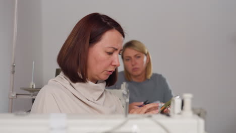 creative women sitting beside sewing machines as one talks while checking smartphone and other listens attentively, focused in textile workshop environment filled with sewing equipment