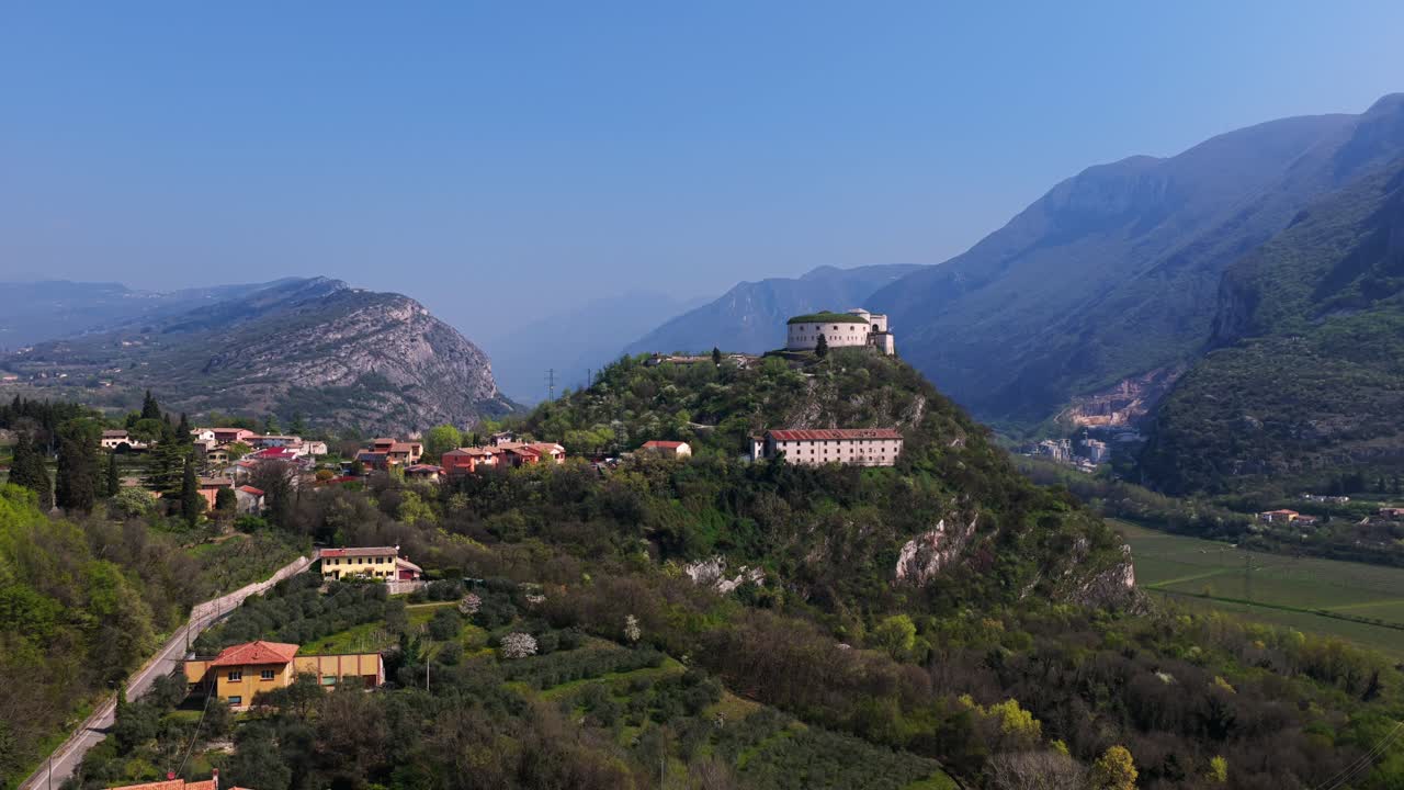 Aerial shot of Rivoli Castle nestled on a hilltop, surrounded by lush greenery and framed by dramatic mountain ranges, capturing the serene beauty of northern Italy's alpine landscape