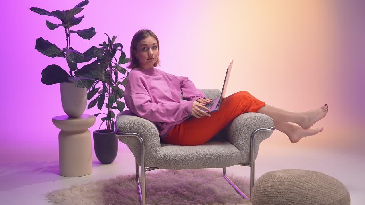 A young woman types on a laptop while lounging barefoot in a soft armchair, surrounded by modern decor, vibrant houseplants, and pastel gradient lighting in a cozy and creative home workspace.