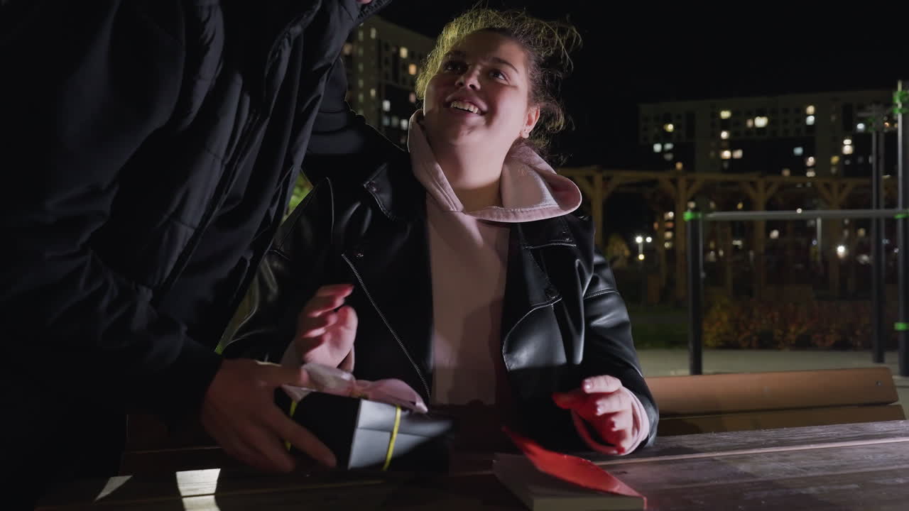 Young girl seated at wooden table reading book during peaceful night scene smiles as her lover presents her with a gift box, they share loving eye contact and tender kiss under soft outdoor lights