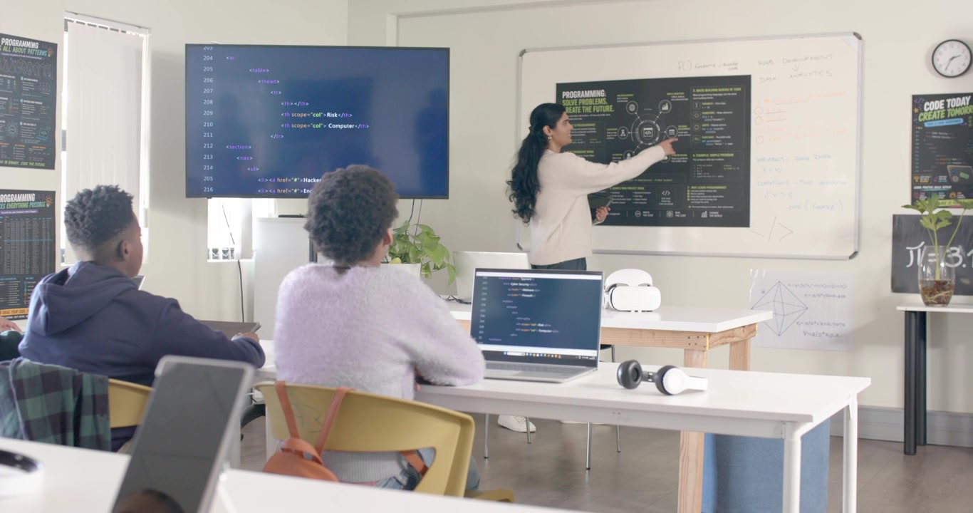 Indian lab teacher pointing at chart, teaching African American teens while screen showing code