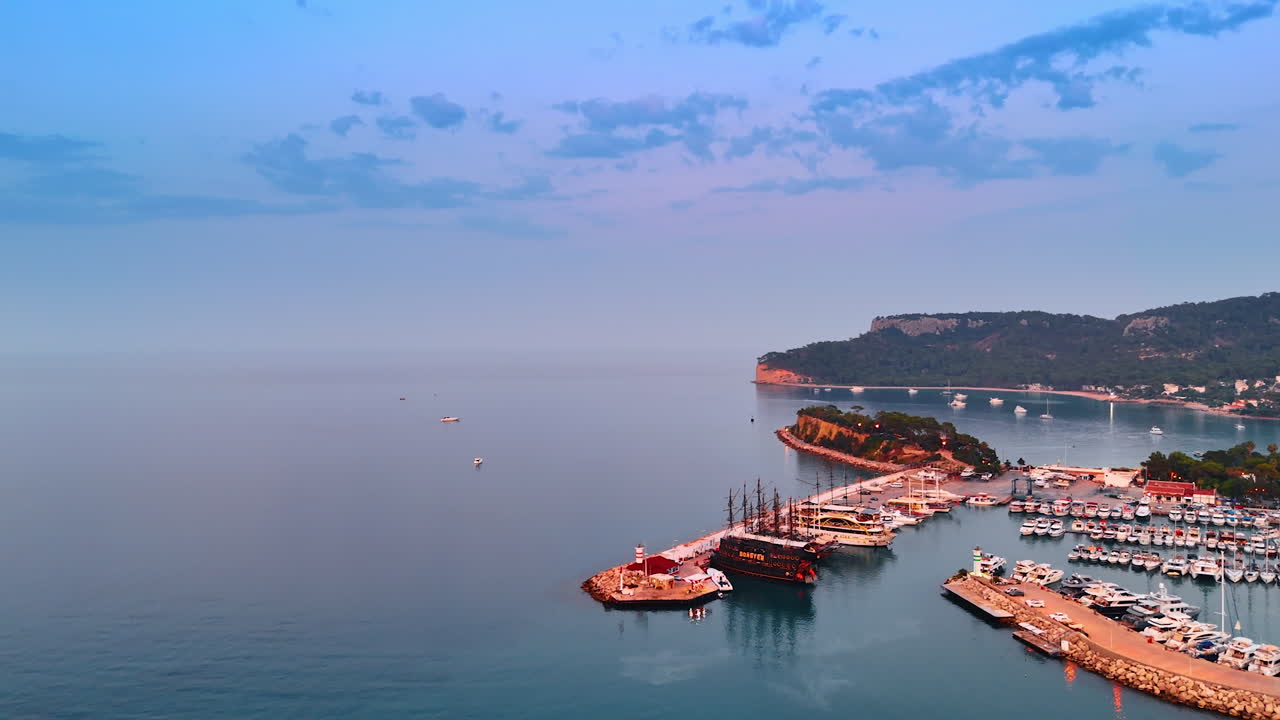 Rising over the calm blue-pink waterscape of the Mediterranean Sea. Flight over the berths with multiple yachts. Kemer, Turkey
