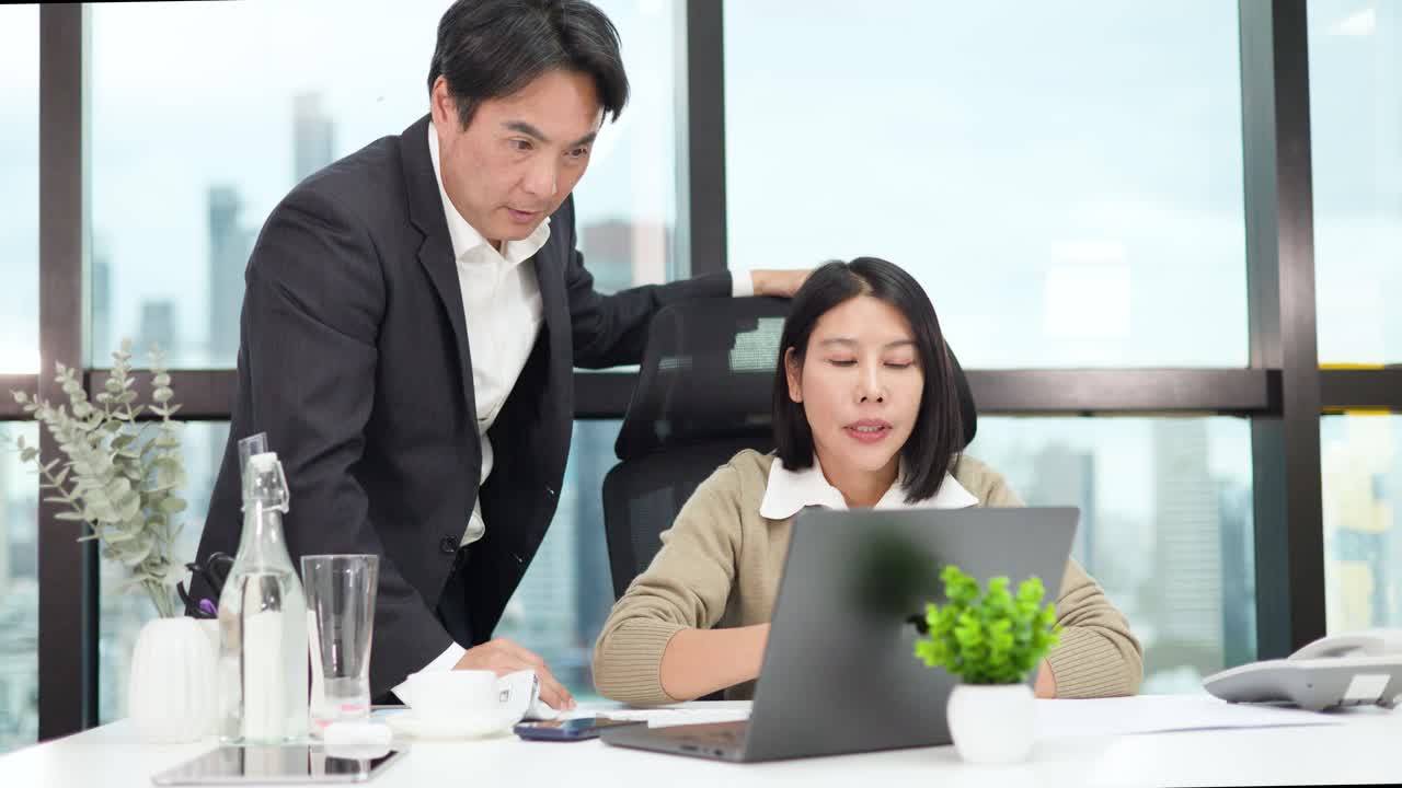 Male manager instructs female employee at desk, pointing at laptop, in bright modern office setting