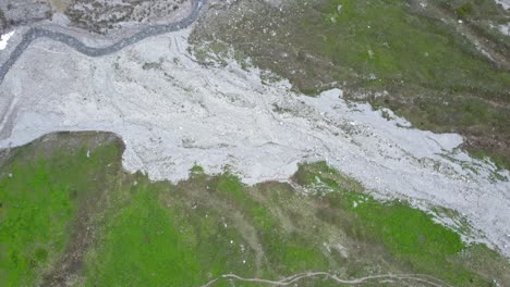 Imágenes-Aéreas-De-Drones-Que-Bajan-Lentamente-En-Espiral-Hacia-Un-Lecho-De-Río-Gris-Ventoso-Y-Un-Prado-Alpino-Verde-Y-Cubierto-De-Hierba-En-Las-Montañas-De-Suiza