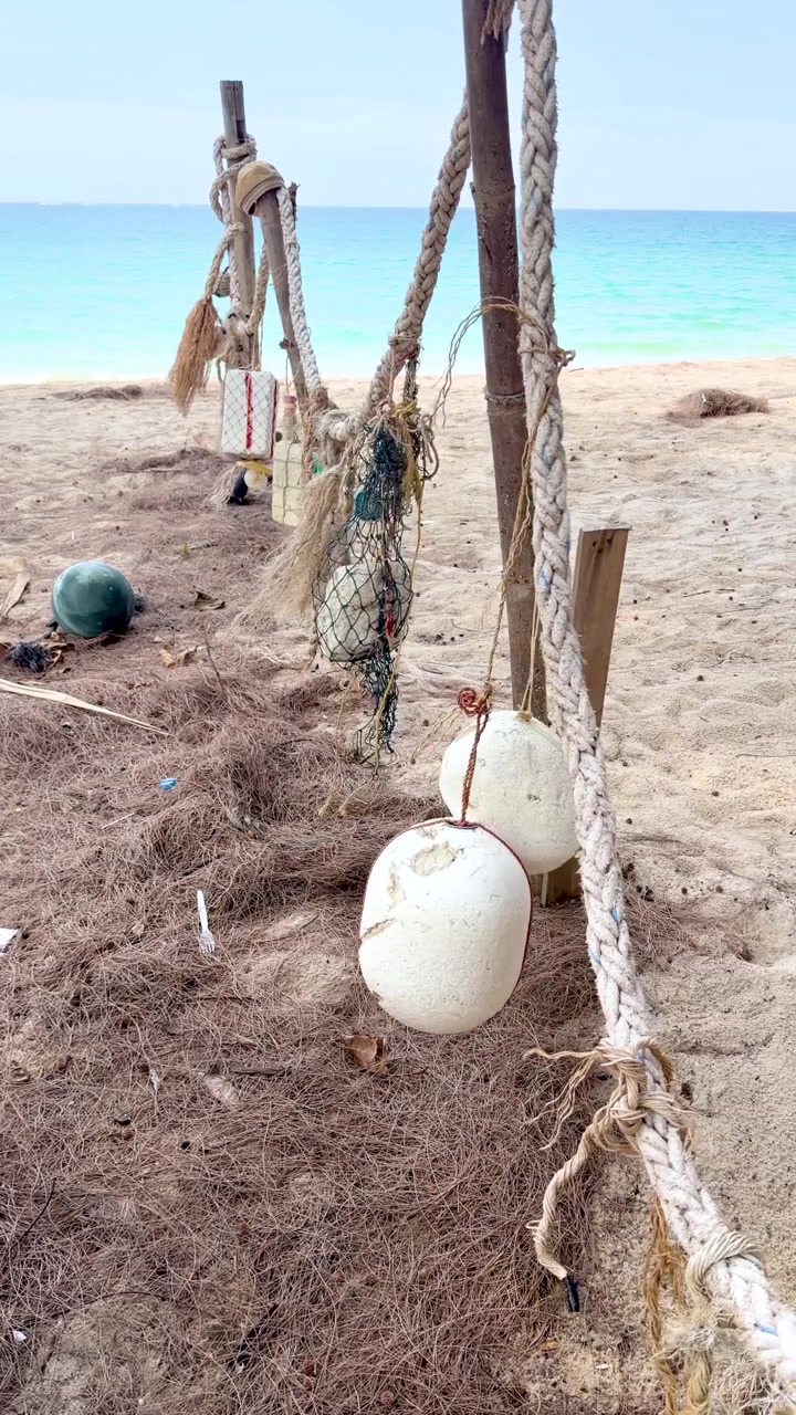 Boe da pesca bianche scartate e corde sfilacciate pendono da pali di legno su una spiaggia sabbiosa.