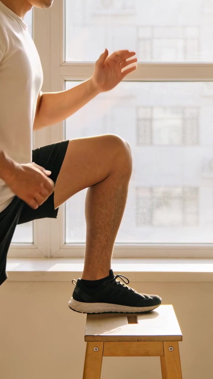 Video verticale: Iniziando il riscaldamento, un uomo in maglietta bianca fa step-up vicino alla finestra usando uno sgabello per il fitness.
