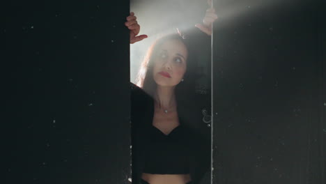 enigmatic woman in black outfit framed by a doorway, softly illuminated by background light as camera zooms in