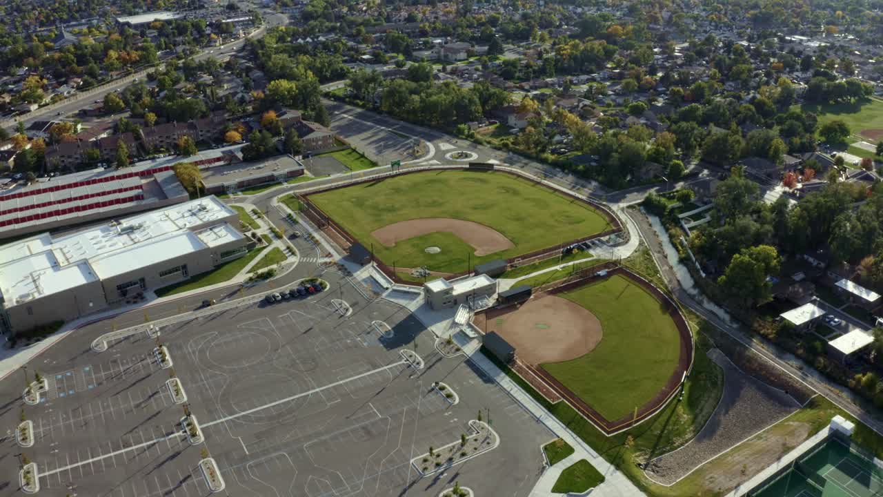 Dolly in aerial drone wide shot approaching empty baseball diamond fields from above behind an American high school surrounded by colorful trees and houses on a sunny fall day in Utah