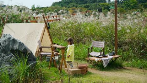 Blond-Child-Girl-at-Summer-Camping-Site-in-Nature-Explore-Kitchen-Utensils-by-Old-Style-Tent,-Ecorium-Botanical-Garden---wide-slow-motion