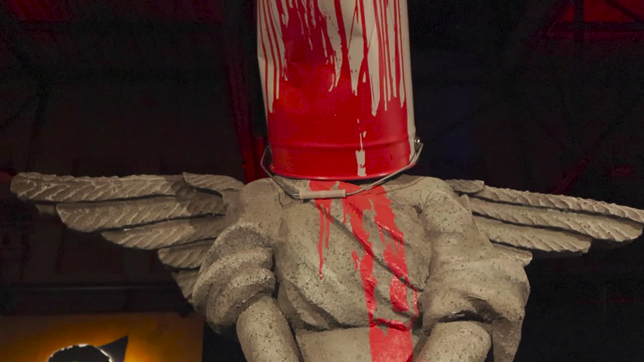 A stone sculpture of a angel with wings who has a red paint bucket on her head done by Banksy at his exhibition in Telliskivi. The statue is well lit and appears to be holding some flowers in her hand