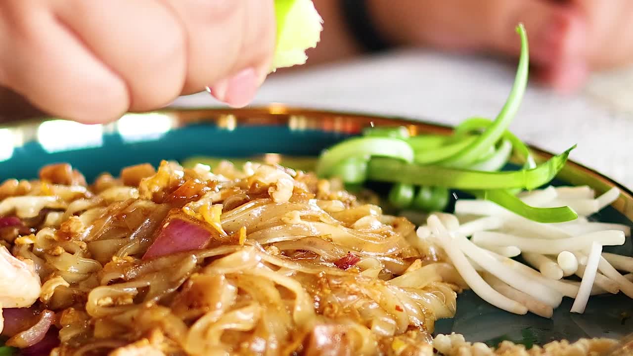 Close-up of hands squeezing lime over a plate of noodles with fresh vegetables.