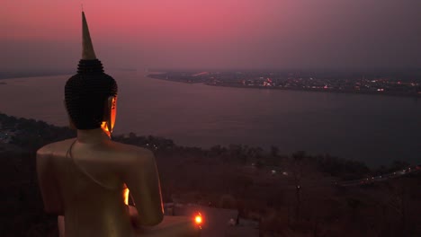 Golden-illuminated-Buddha-overlooking-Mekong-river-vibrant-sunset,-Pakse,-Laos