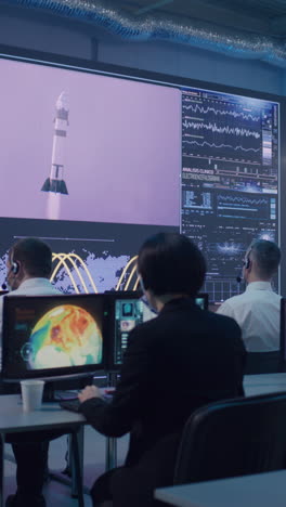 Boss-and-employees-watching-successful-launch-of-rocket-on-large-screen-then-applauding-and-giving-high-five-during-work-in-flight-control-center.-Vertical-shot