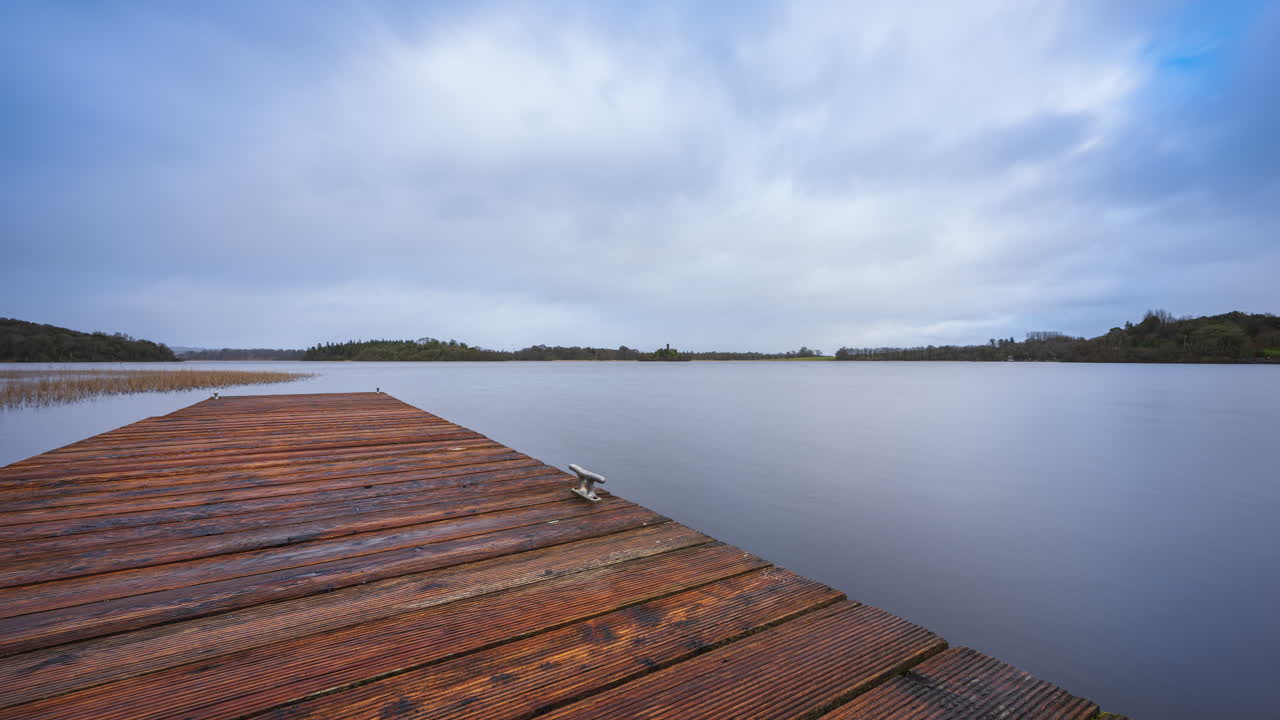 Time lapse of a timber lake pier in the foreground and castle ruin island with forest in distance on a cloudy sunny day at Lough Key in county Roscommon in Ireland