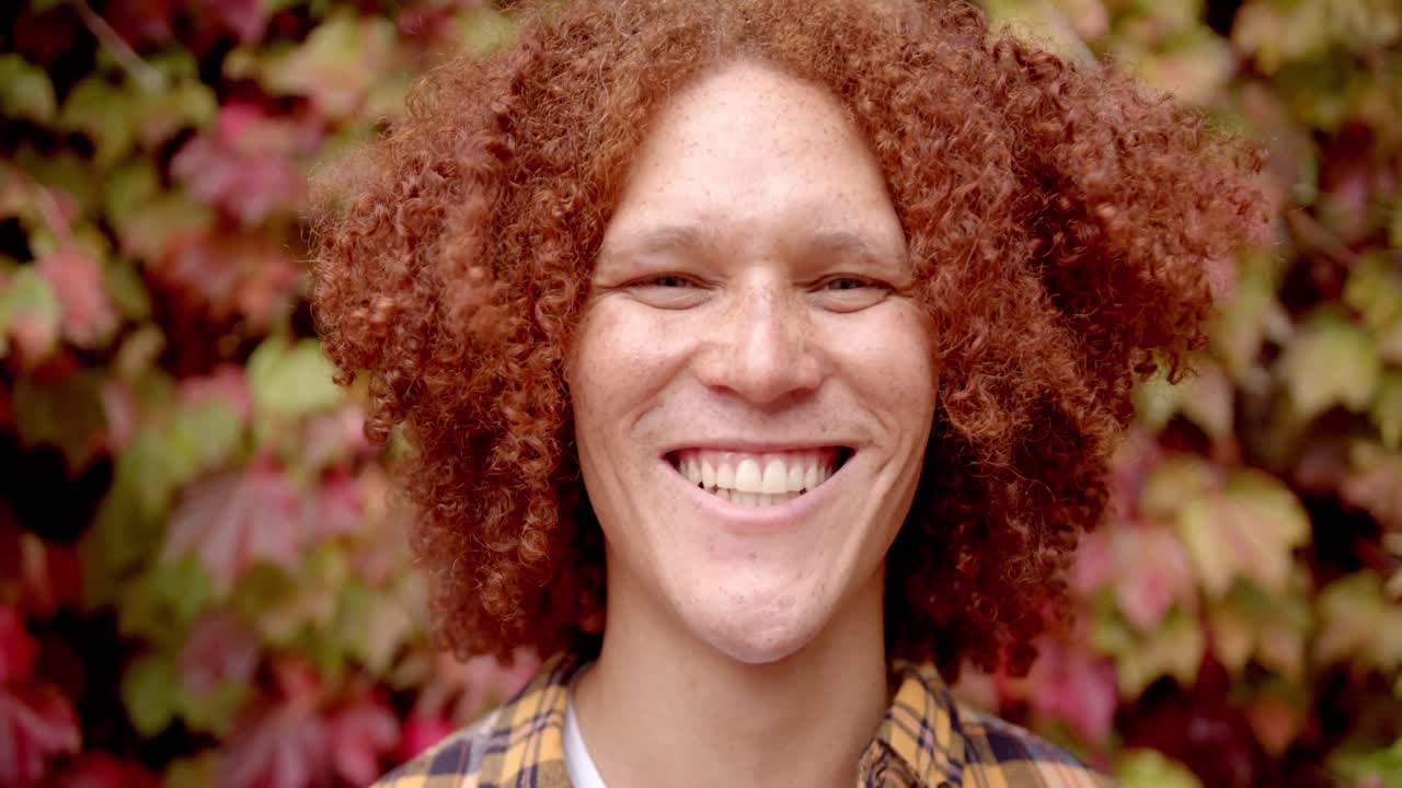 retrato de un feliz hombre biracial con cabello rojo rizado y pecas sonriendo en el jardín, cámara lenta