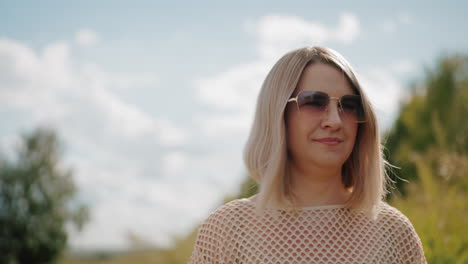 business lady in net cloth taking stroll with warm smile, wearing sunglasses under sunny sky, with blurred background featuring lush greenery