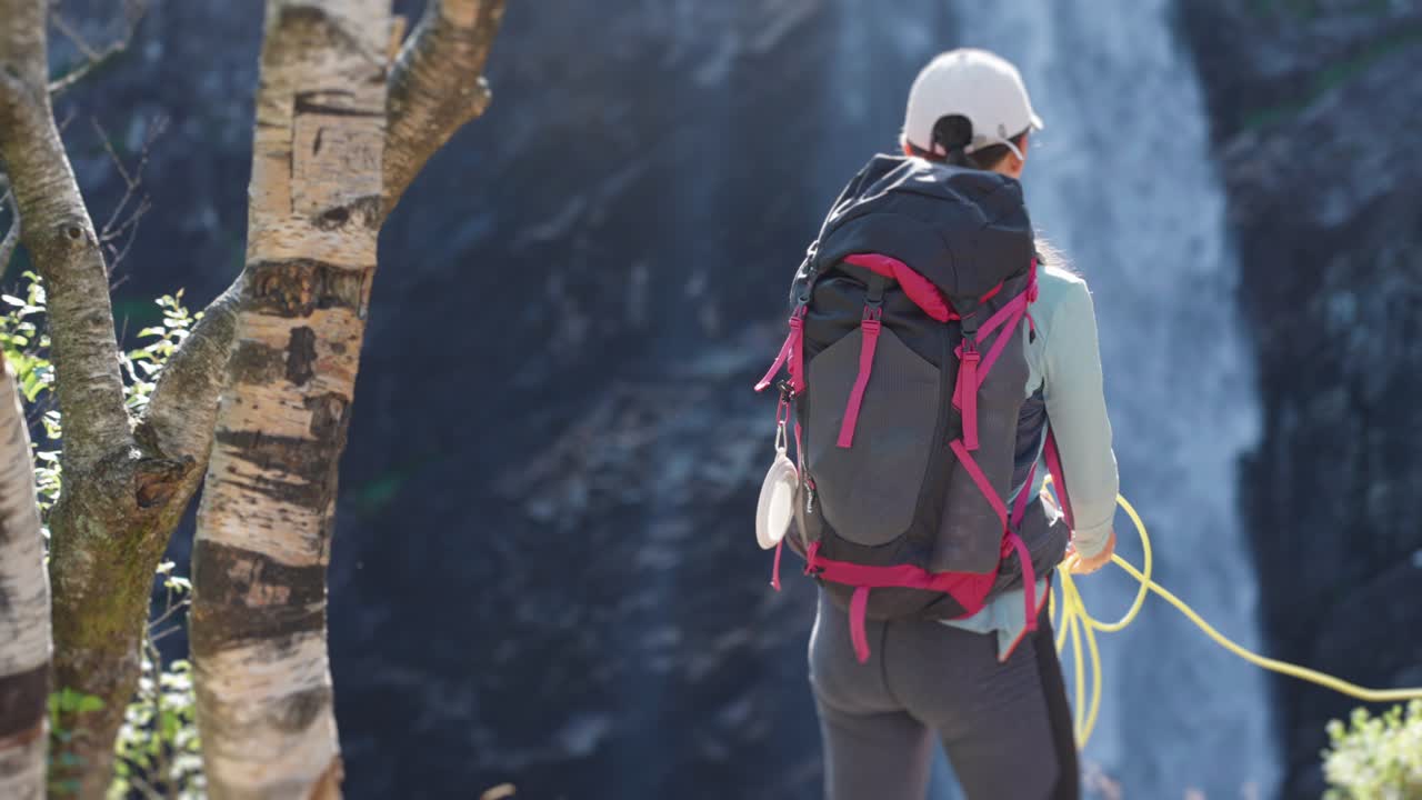 Una vista posteriore dell'escursionista donna con un guinzaglio tra le mani, in piedi sulla piattaforma mentre osserva la cascata.