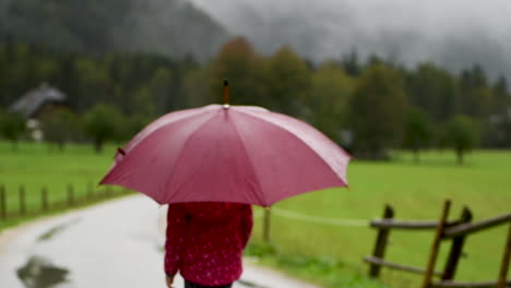 Niña-Caminando-Juguetonamente-Bajo-La-Lluvia-En-Una-Carretera-Rural,-Una-Granja-En-El-Fondo,-Un-Valle-Alpino,-Cerca-Desde-Atrás,-Desenfoque-Intencional,-Desde-Atrás-Mirando-Hacia-Afuera