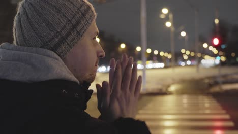 side portrait of young man warming up hands with breath, cold winter city night