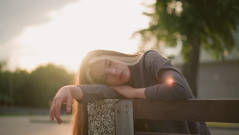 lady seated at edge of bench, resting head on arm, surrounded by serene outdoor park setting with soft sunlight reflecting around her, background featuring tree and building