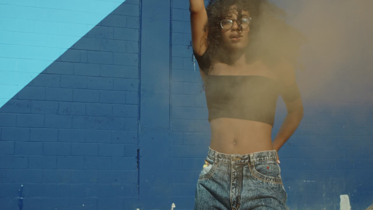 hermosa mujer joven sosteniendo colorida granada de humo bailando afuera contra la pared azul