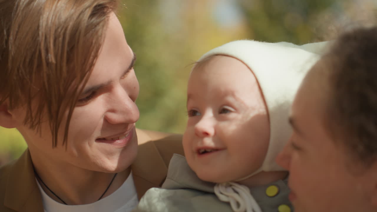 Nærbillede af kaukasisk ægtepar i park, der morer spædbarnet med sjove ansigter og blide pust. Baby peger på fars næse, mens forældrene griner, kærlig interaktion, oprigtige smil, hyggeligt familiemoment.