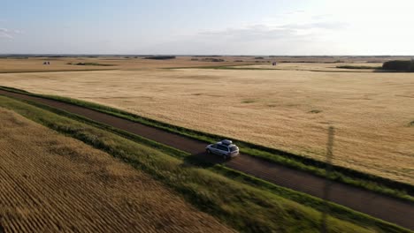 4K-Luftaufnahme-Eines-Silbernen-Autos,-Das-Bei-Sonnenuntergang-In-Der-Prärie-Auf-Einer-Staubigen-Landstraße-Fährt