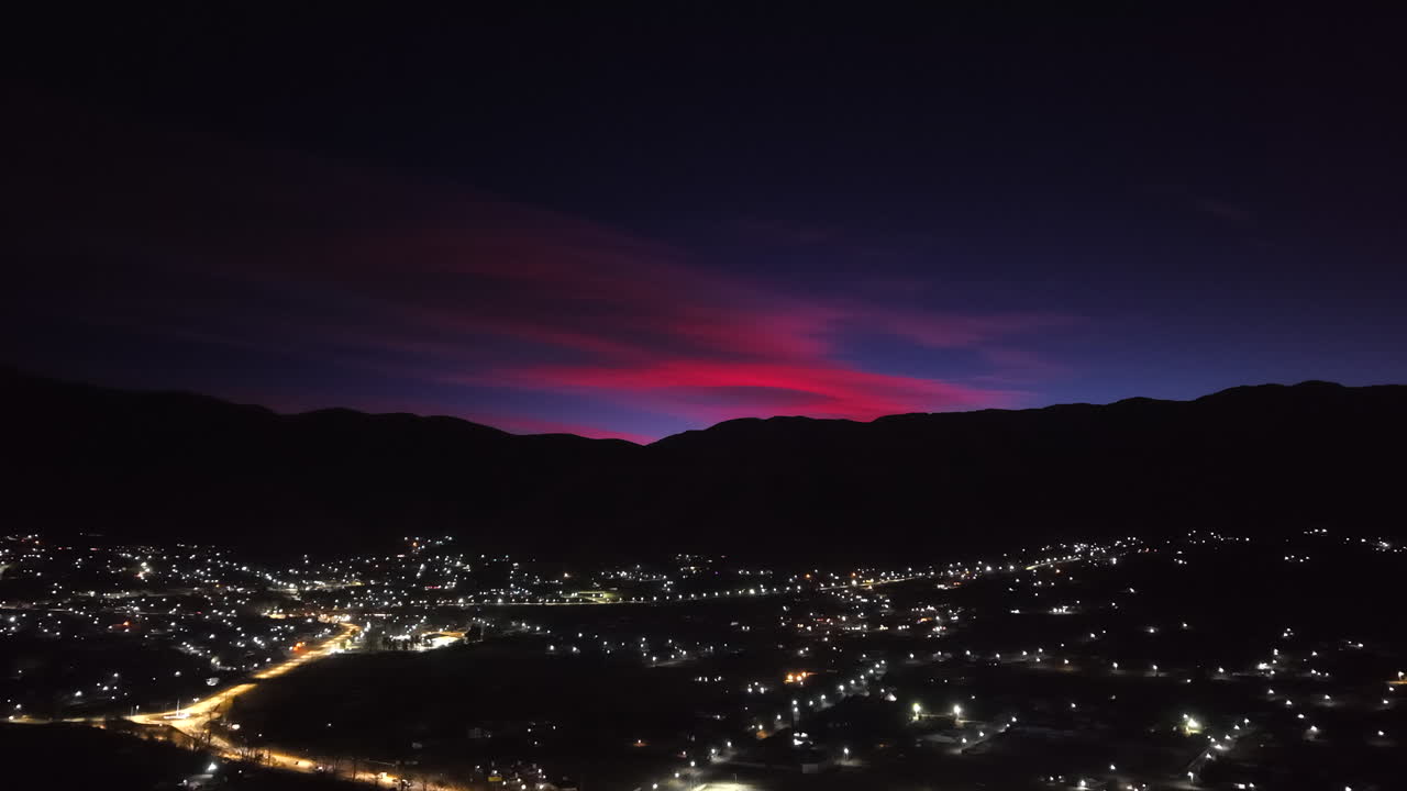 A stunning aerial shot pulls back and rises to reveal a beautiful sunrise painting the sky red above the illuminated mountain town of Tafí del Valle in the Andes of Argentina