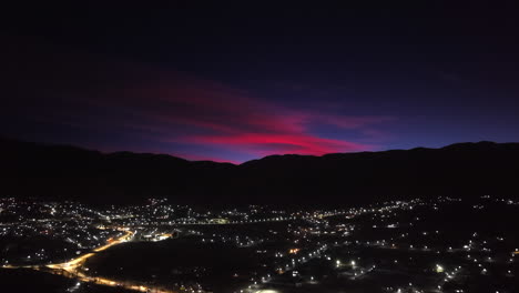 A stunning aerial shot pulls back and rises to reveal a beautiful sunrise painting the sky red above the illuminated mountain town of Tafí del Valle in the Andes of Argentina