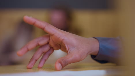 close-up of young man's hand hovering above wooden table in relaxed, curved position with slight tension in fingers, suggesting anticipation or gesture, soft indoor background with blurred human figures