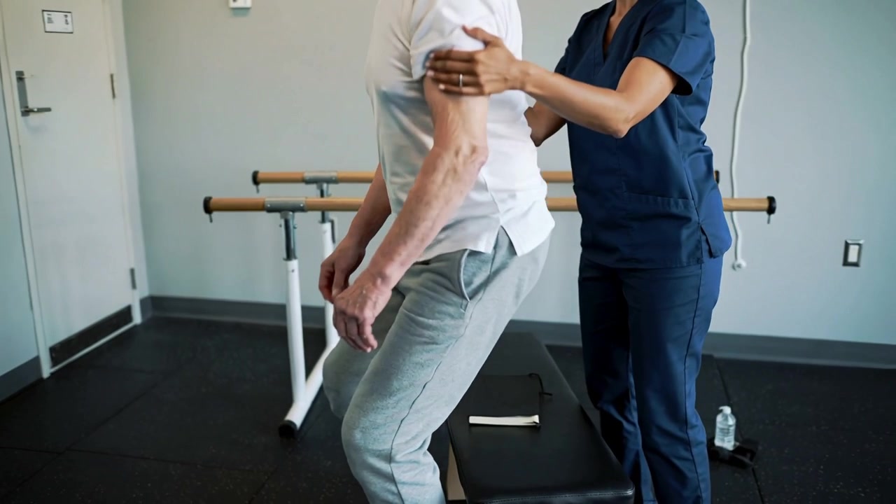 Eldre mann lener seg og skyver seg fra en benk mens han øver på å stå, med hjelp fra ansatte på en klinikk, ved støttestenger.