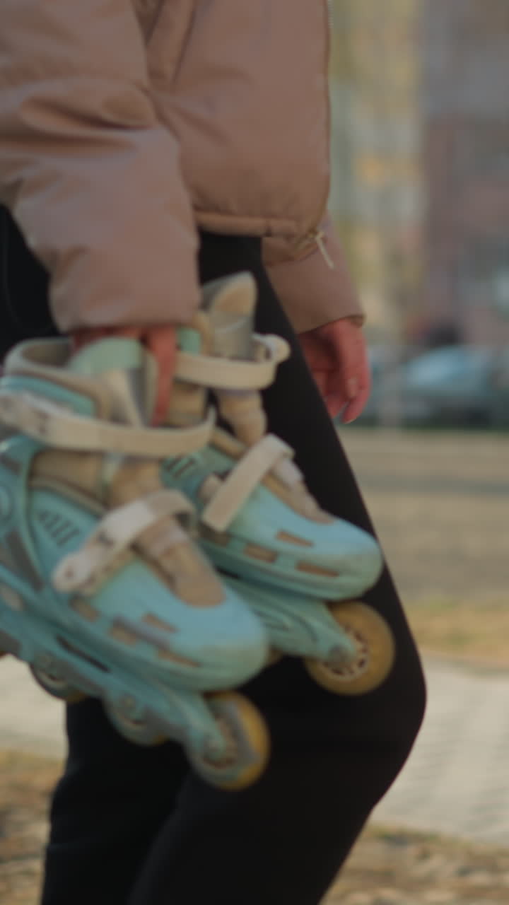 얼굴이 없는 사람이 한 손에 롤러블레이드를 들고 빛이 비치는 공원을 걸어가고 있다. 개인은 복아 재과 검은 바지를 입고 있으며, 배경에는 흐릿한 집들이 있다.