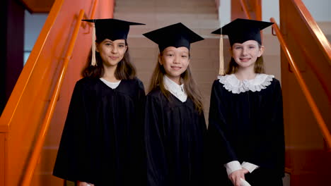 zoom out of three happy preschool female students in cap and gown posing and looking at the camera at the graduation ceremony