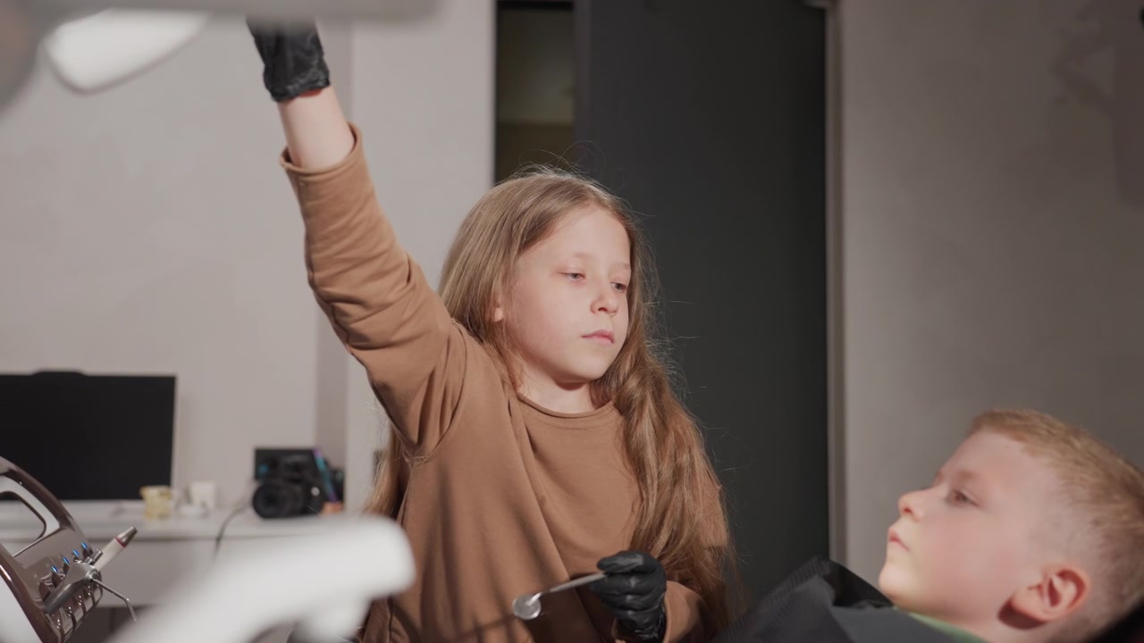 Close up of young girl wearing black gloves holding dental mirror and instrument while examining boy in dentist chair. Dental equipment including light monitor visible on left side. Indoor