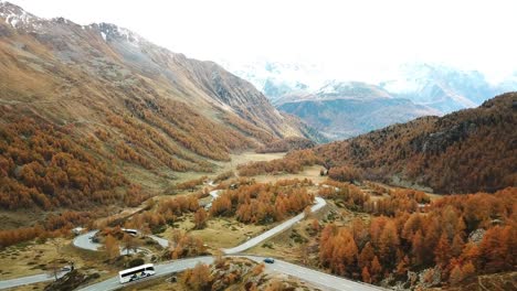 Aerial view of a winding road through a mountainous landscape with autumn-colored trees, surrounded by rugged peaks and a partly cloudy sky, capturing the serene beauty of nature from above