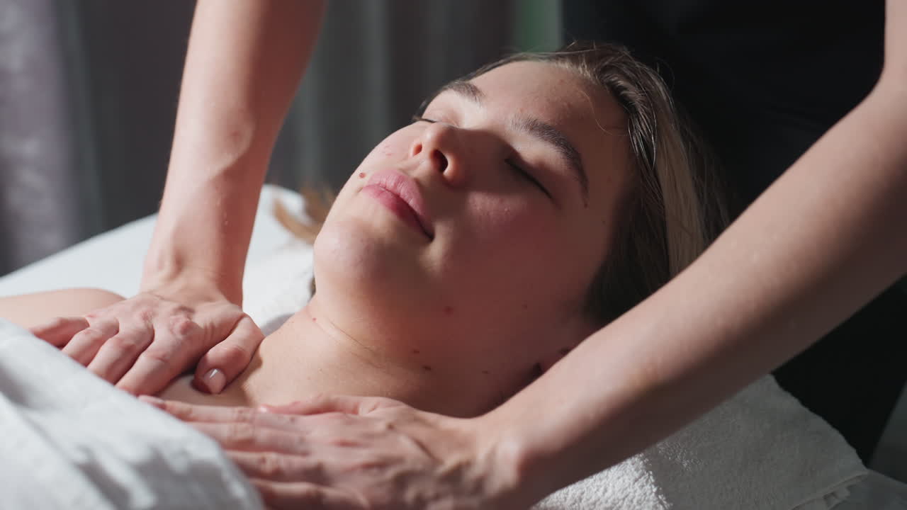 Massage therapist in black uniform gently massaging collarbone and upper chest of light-skinned female client lying with eyes closed on spa bed, showing peaceful relaxation under soft natural light