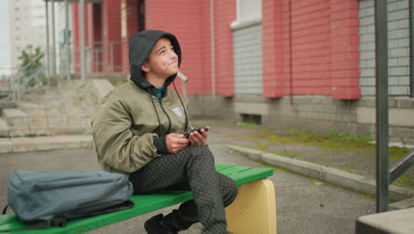 boy in hooded jacket seated on green bench holding phone, glancing up with subtle smile, gray backpack beside him, steps and brick wall in background, relaxed outdoor urban moment
