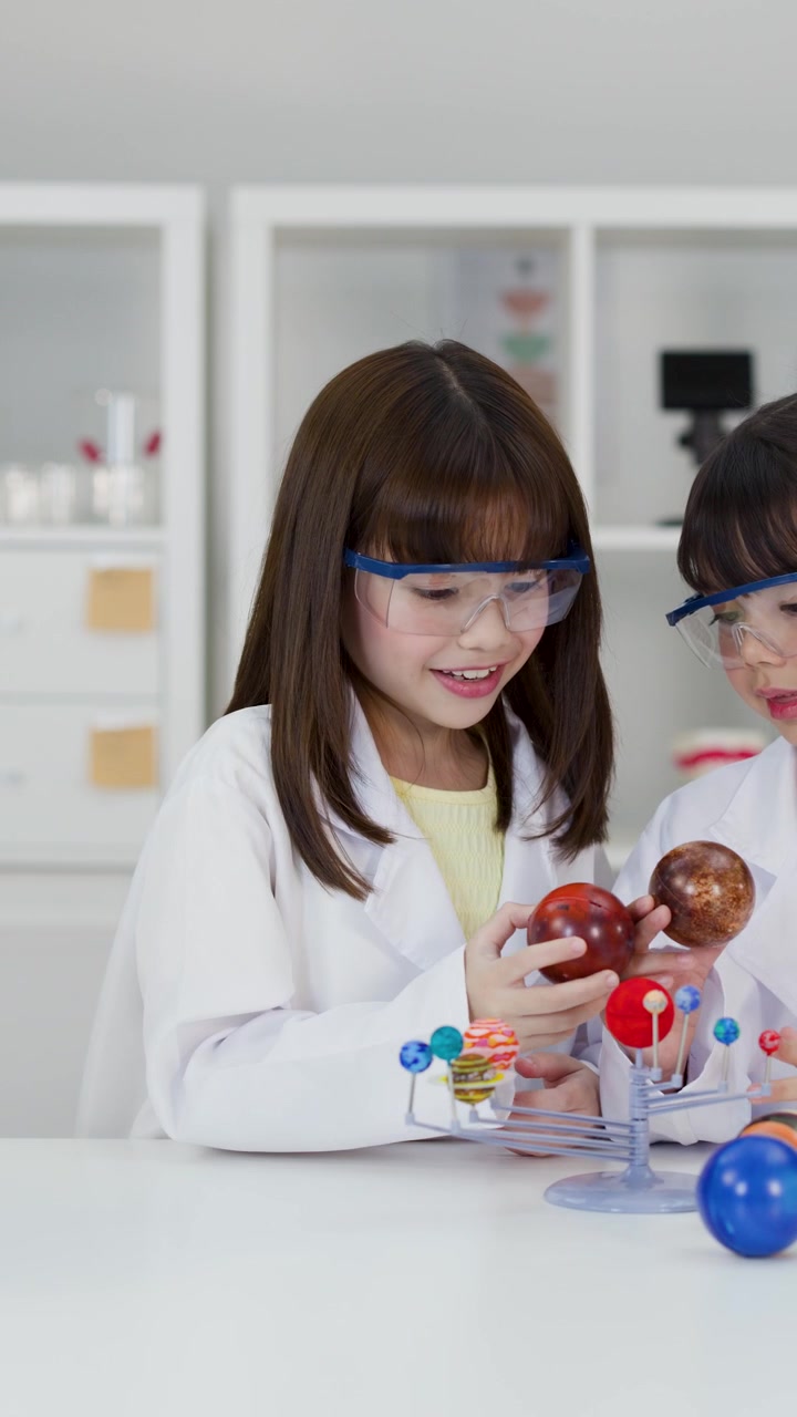 Young students in lab coats and goggles examine a planetary model in a bright classroom.